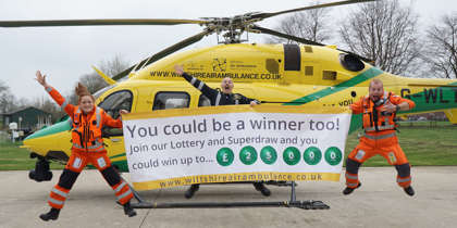 Two paramedics and a pilot jumping and smiling in front of the helicopter on a helipad. They are holding a large white and yellow banner advertising the Lottery Superdraw. Two paramedics and a pilot jumping and smiling in front of the helicopter on a helipad. They are holding a large white and yellow banner advertising the Lottery Superdraw.