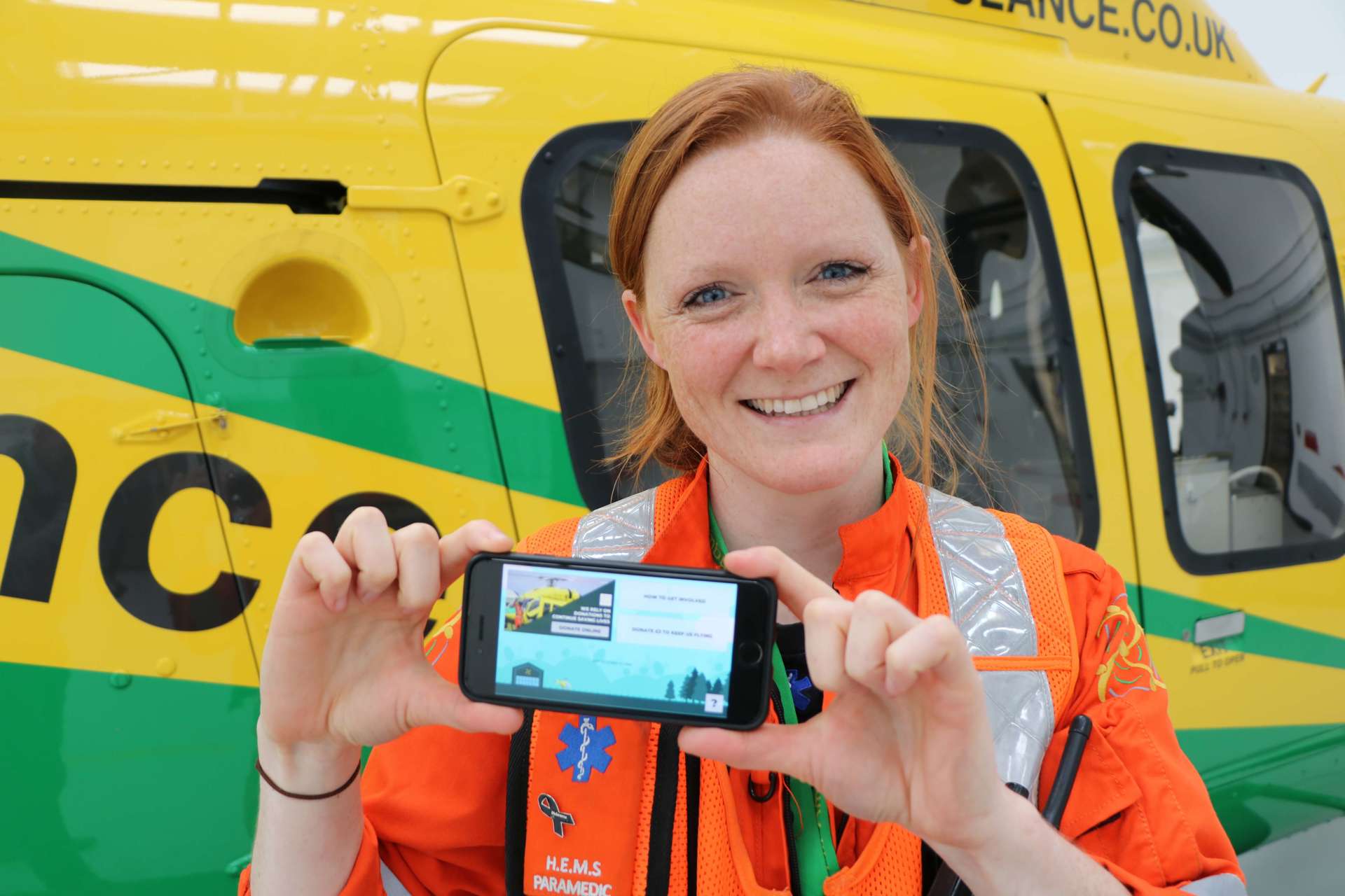 A critical care paramedic stood holding a phone with the app Helifun on screen. A critical care paramedic stood holding a phone with the app Helifun on screen.