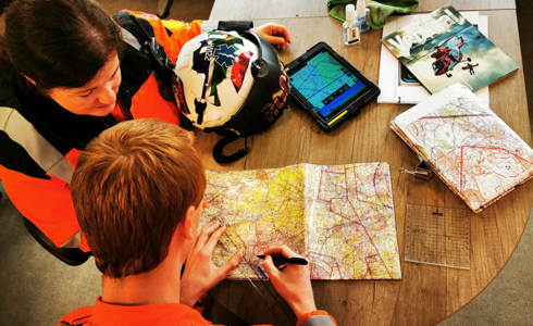 Two paramedics who are both wearing orange flight suits. They are both sat at a wooden desk with an iPad and paper maps and are plotting a route.