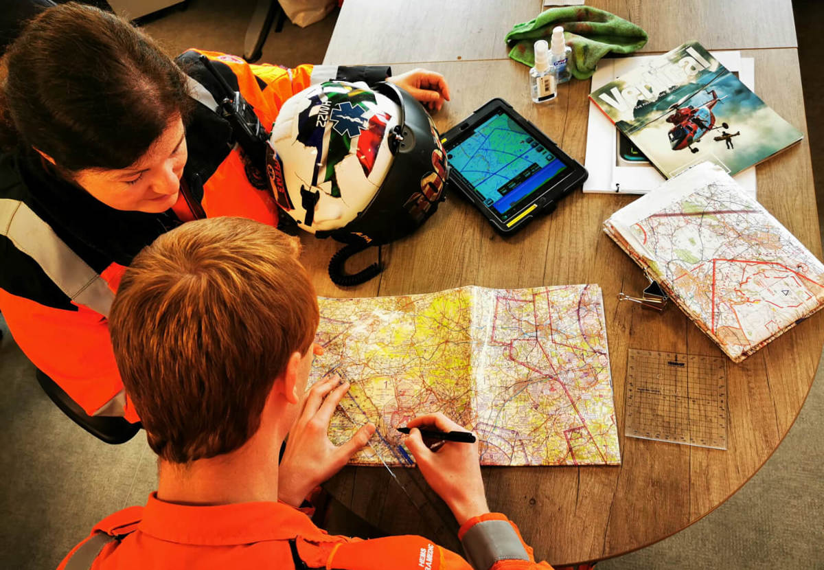 Two paramedics who are both wearing orange flight suits. They are both sat at a wooden desk with an iPad and paper maps and are plotting a route.