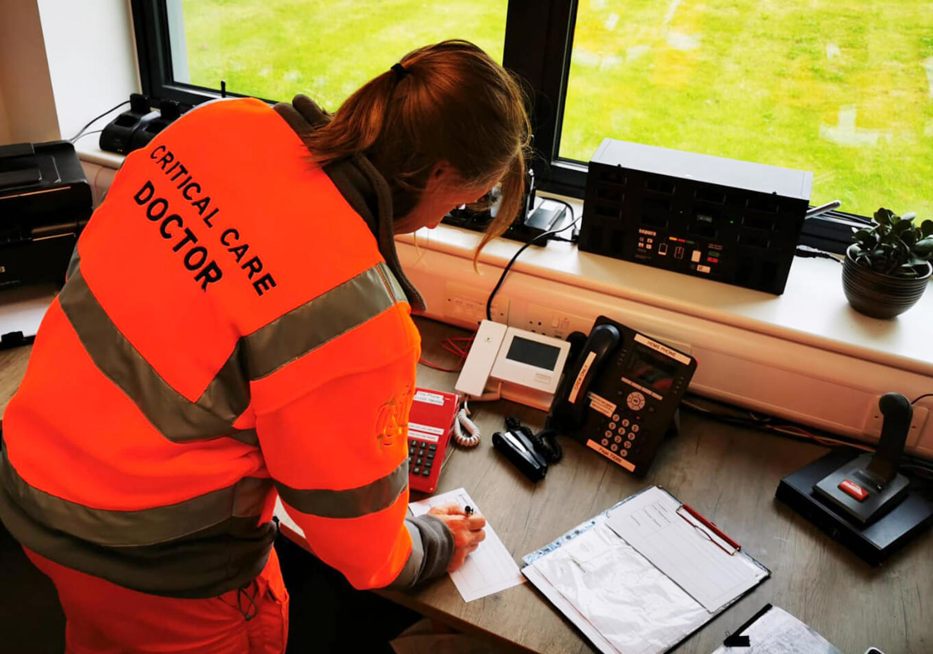 A doctor wearing an orange fleece taking notes whilst on a 999 call whilst stood at a wooden desk with paperwork and a clipboard. A doctor wearing an orange fleece taking notes whilst on a 999 call whilst stood at a wooden desk with paperwork and a clipboard.