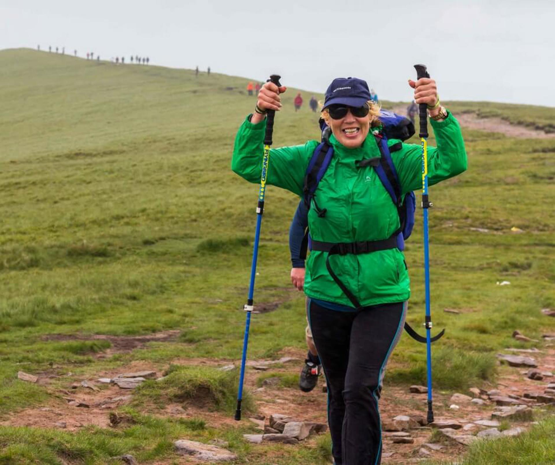 A person wearing a green raincoat and blue backpack, holding walking poles in the air