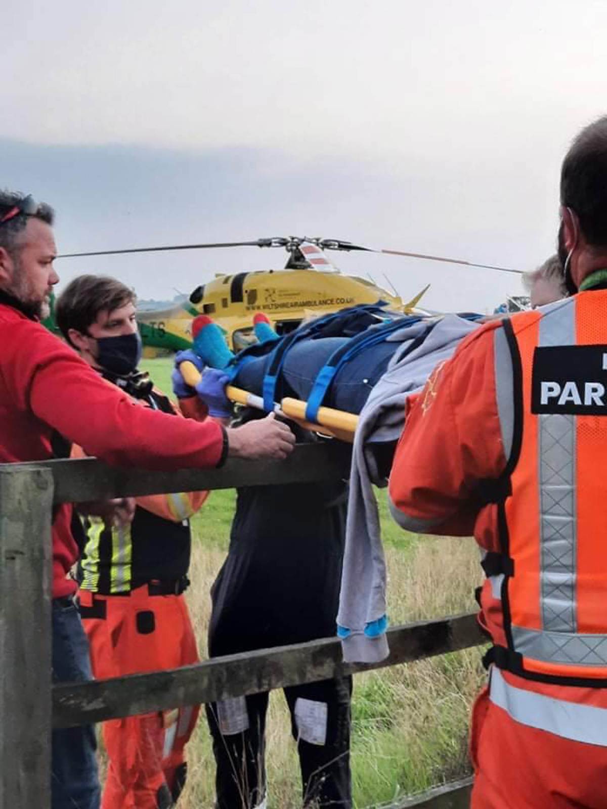 A patient being carried over a fence on a stretcher. The Bell 429 helicopter is in the background. A patient being carried over a fence on a stretcher. The Bell 429 helicopter is in the background.