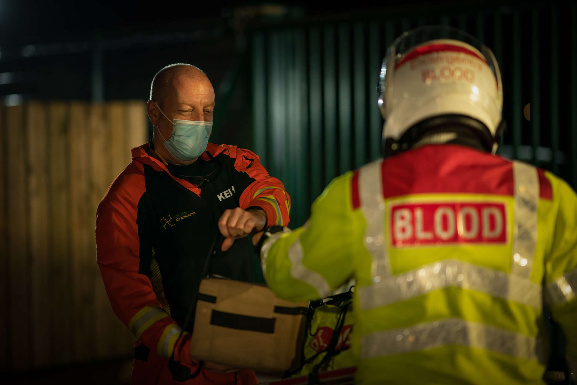 A paramedic who is being handed a delivery box of blood products from a biker wearing a high vis jacket and a helmet. A paramedic who is being handed a delivery box of blood products from a biker wearing a high vis jacket and a helmet.