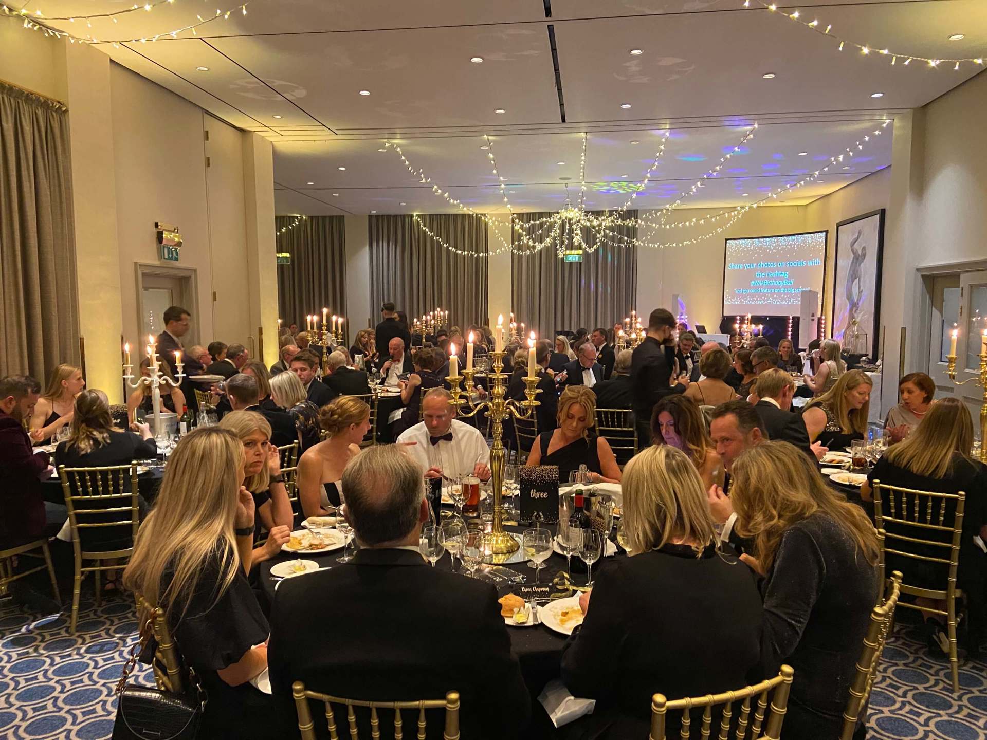 Black and gold themed charity ball. Groups of people wearing smart clothing, sitting on gold chairs around round tables Black and gold themed charity ball. Groups of people wearing smart clothing, sitting on gold chairs around round tables