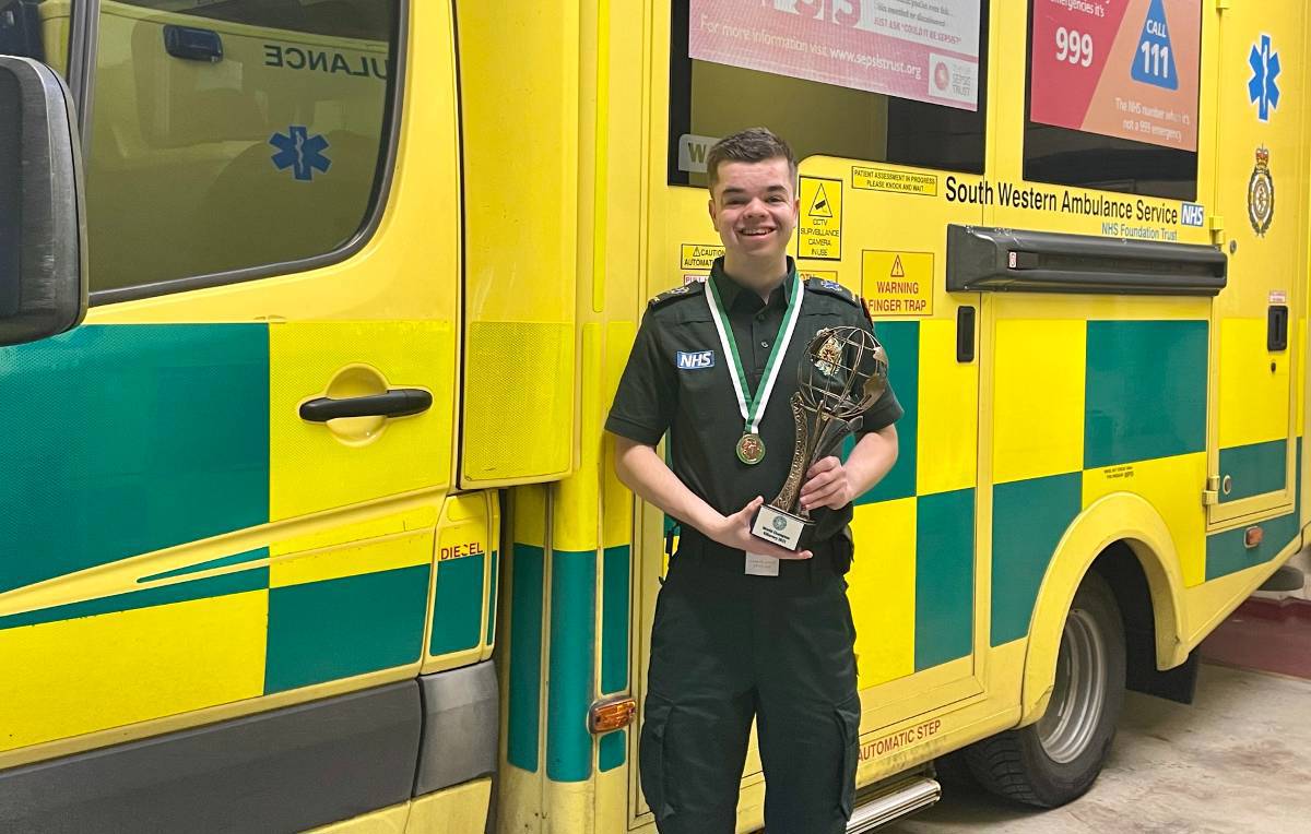 Leon in uniform holding the World Irish Dance Champion trophy, with a South Western Ambulance Service NHS Foundation Trust vehicle