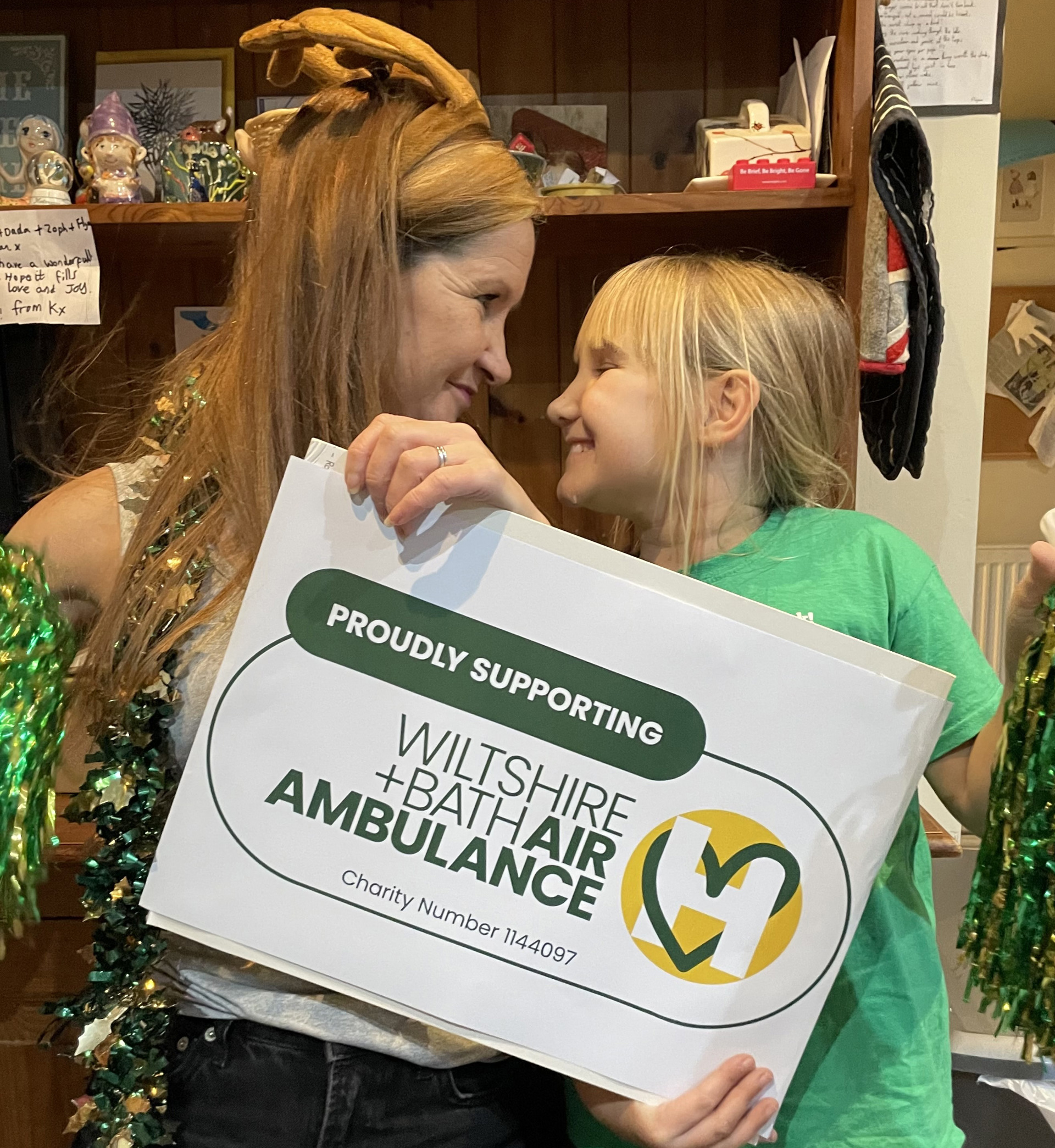 A lady and girl holding a proudly supporting Wiltshire and Bath Air Ambulance logo, whilst wearing greeb