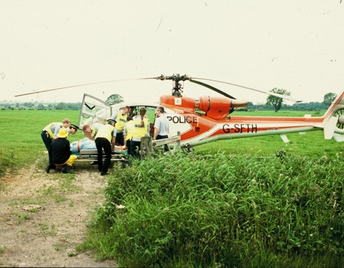 A scanned photo of a red helicopter landed in a field with police lifting a patient on a stretcher.