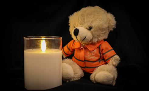 A photo of a lit candle with a small teddy bear sat next to it, wearing an orange flight suit, on a black background