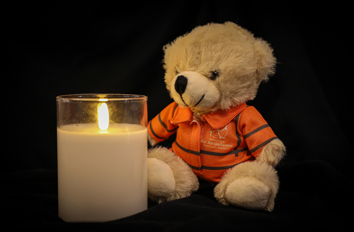A photo of a lit candle with a small teddy bear sat next to it, wearing an orange flight suit, on a black background A photo of a lit candle with a small teddy bear sat next to it, wearing an orange flight suit, on a black background