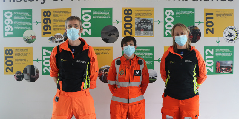 Three doctors stood in front of a history timeline wearing blue face coverings.