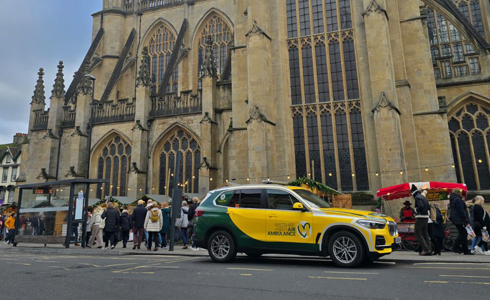 A Wiltshire and Bath Air Ambulance Charity branded green and yellow BMW X5 parked outside a busy Bath Abbey with Christmas Market chalets