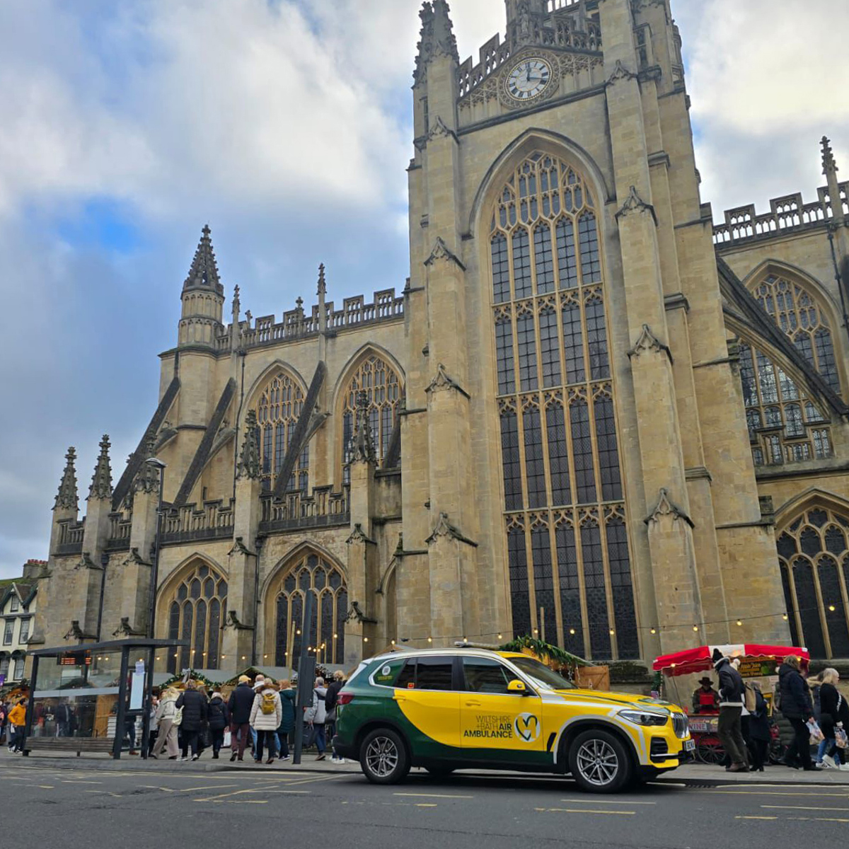 A Wiltshire and Bath Air Ambulance Charity branded green and yellow BMW X5 parked outside a busy Bath Abbey with Christmas Market chalets